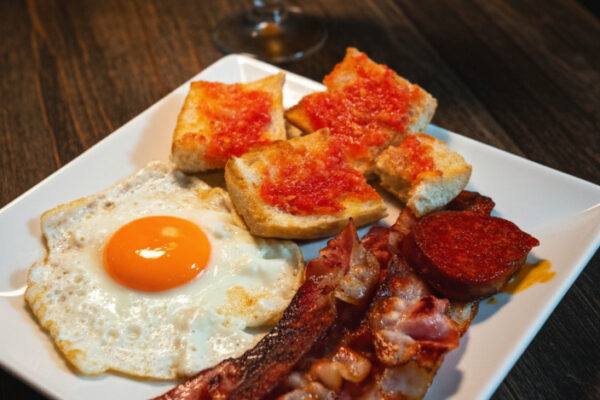 Plato de desayuno o almuerzo fuerte con huevo frito, tiras de beicon crujiente, rodajas de chorizo, y pan con tomate, acompañado de una copa de cerveza oscura en el restaurante Amazonia.